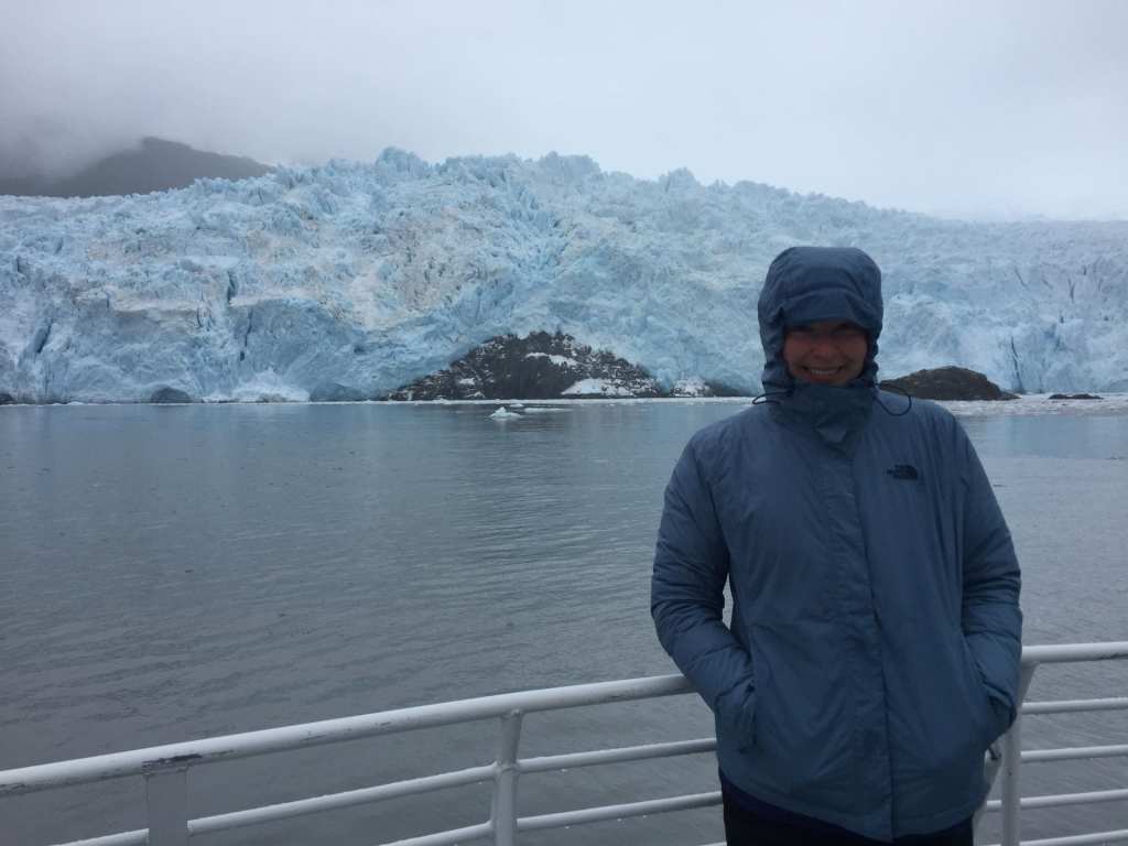 Light blue, heavily crevassed tidewater glacier sitting in dark gray, smooth water under a foggy sky. Person in raincoat leaning on boat railing in foreground.
