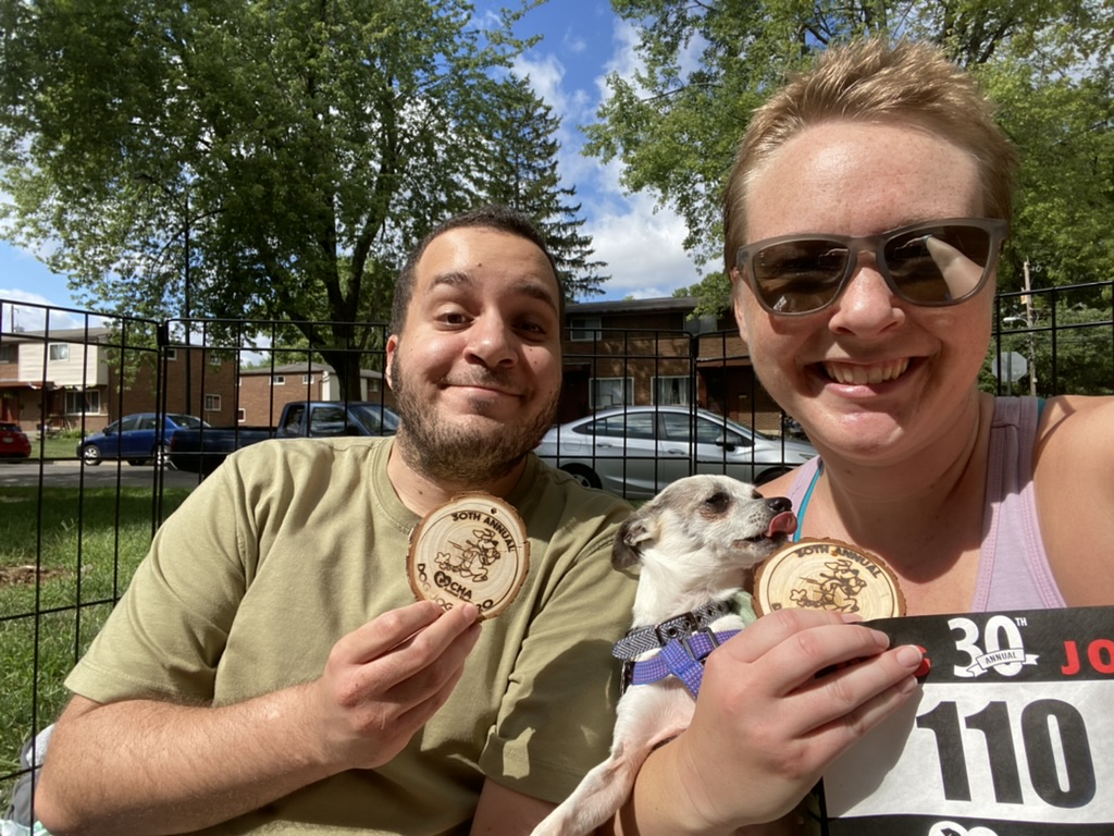 Man in green shirt holding a race medal next to woman in sunglasses holding a white chihuahua mix, a race medal, and a race bib.